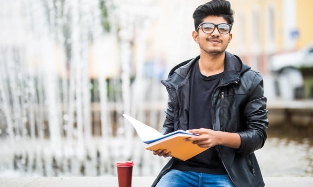 indian student man holding pile books sitting near fountain street 231208 2778