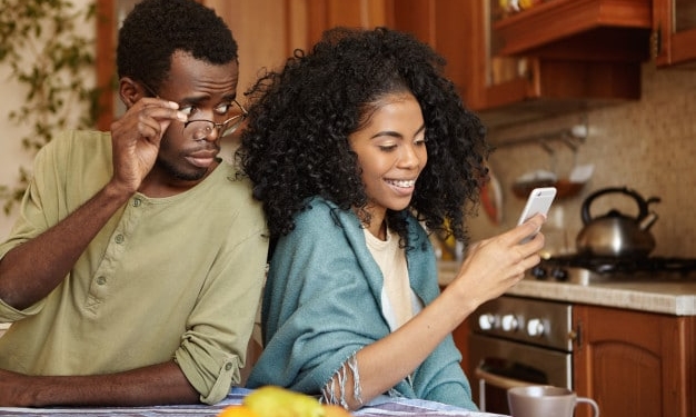 jealous curious black man holding glasses spying his girlfriend s mobile phone while she is typing message her lover smiling happily betrayal unfaithfulness infidelity lack trust 273609 298